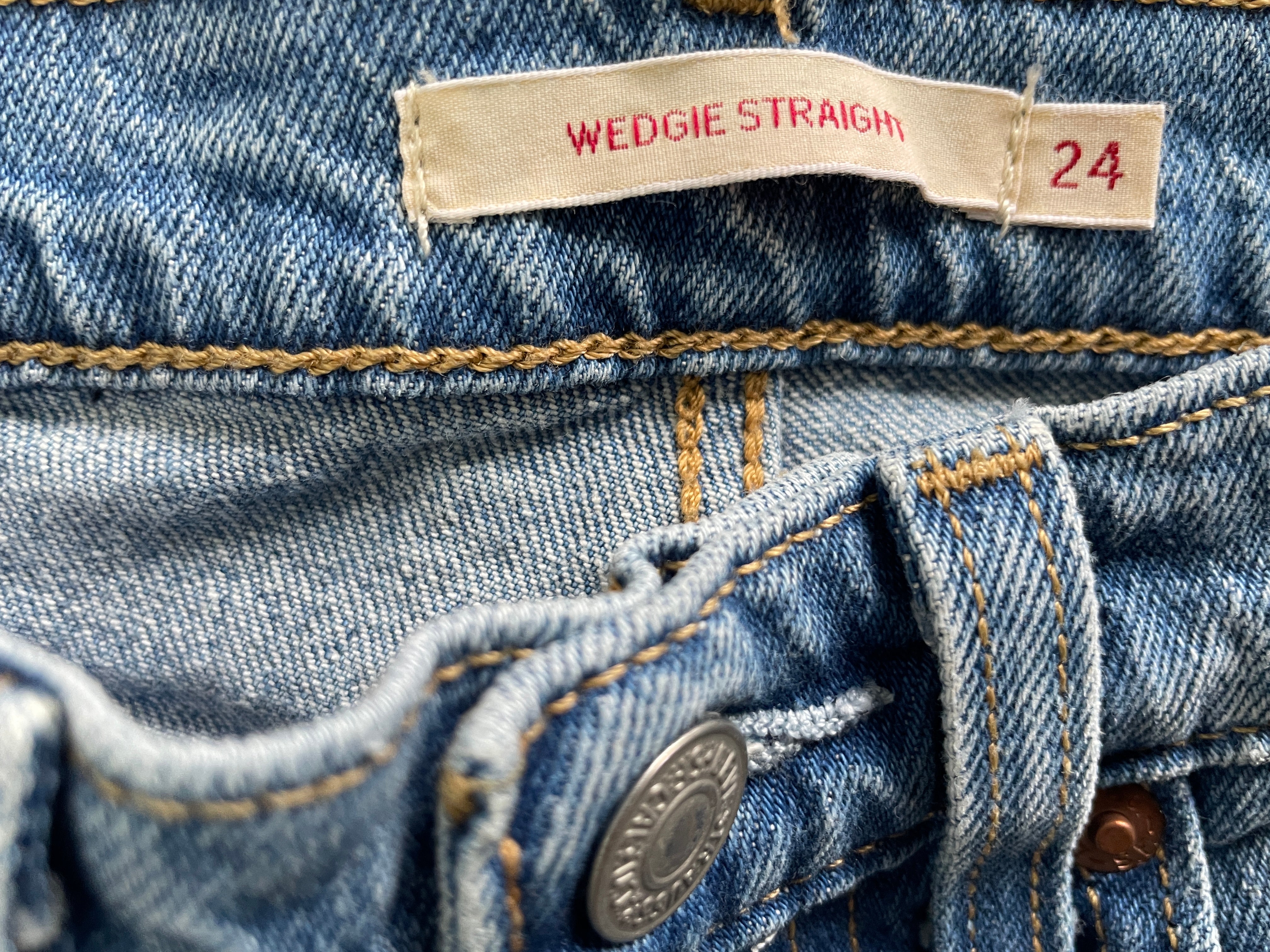 Close-up of a pair of blue denim jeans showing the waistband, a beige fabric label that reads 'WEDGIE STRAIGHT' and '24', stitching, and a metal button.