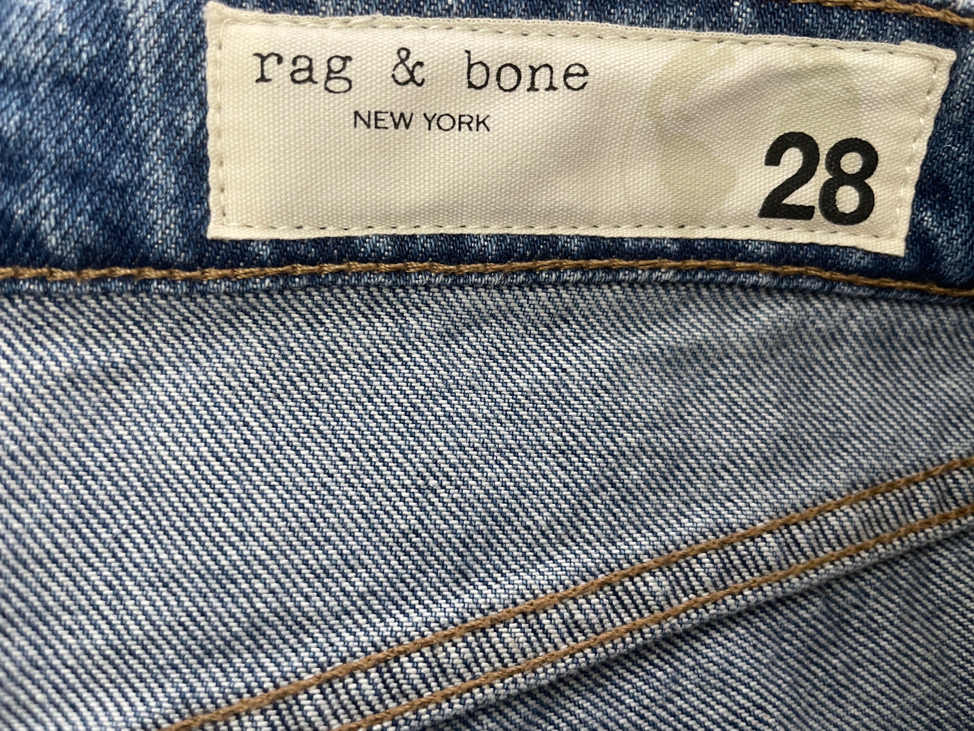 Close-up of a denim garment showing the inner waistband label with the brand name 'rag & bone' and size '28'.