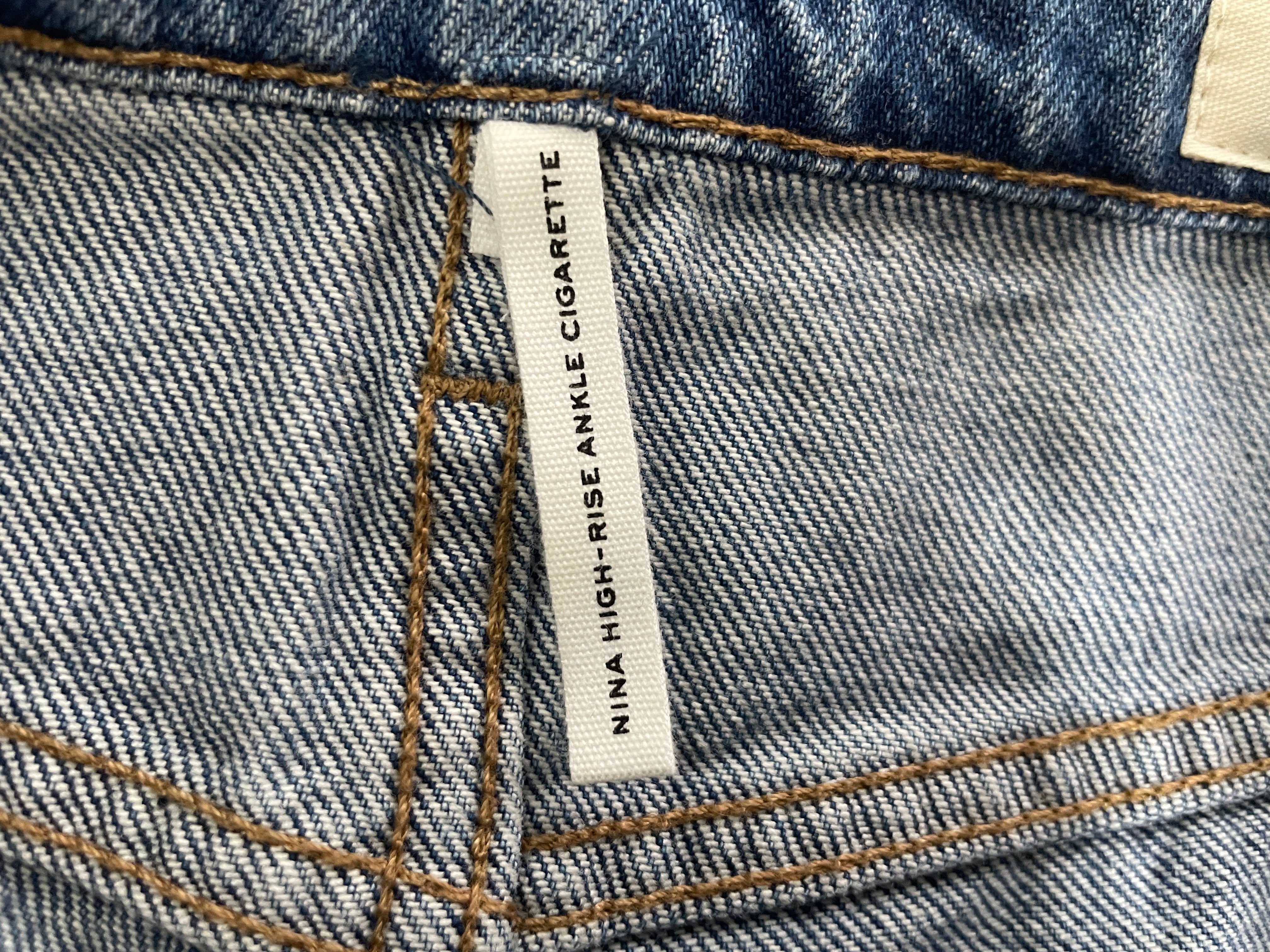 Close-up of a denim waistband showing a white fabric label that reads 'NINA HIGH-RISE ANKLE CIGARETTE' with blue-gray twill fabric and tan contrast stitching.