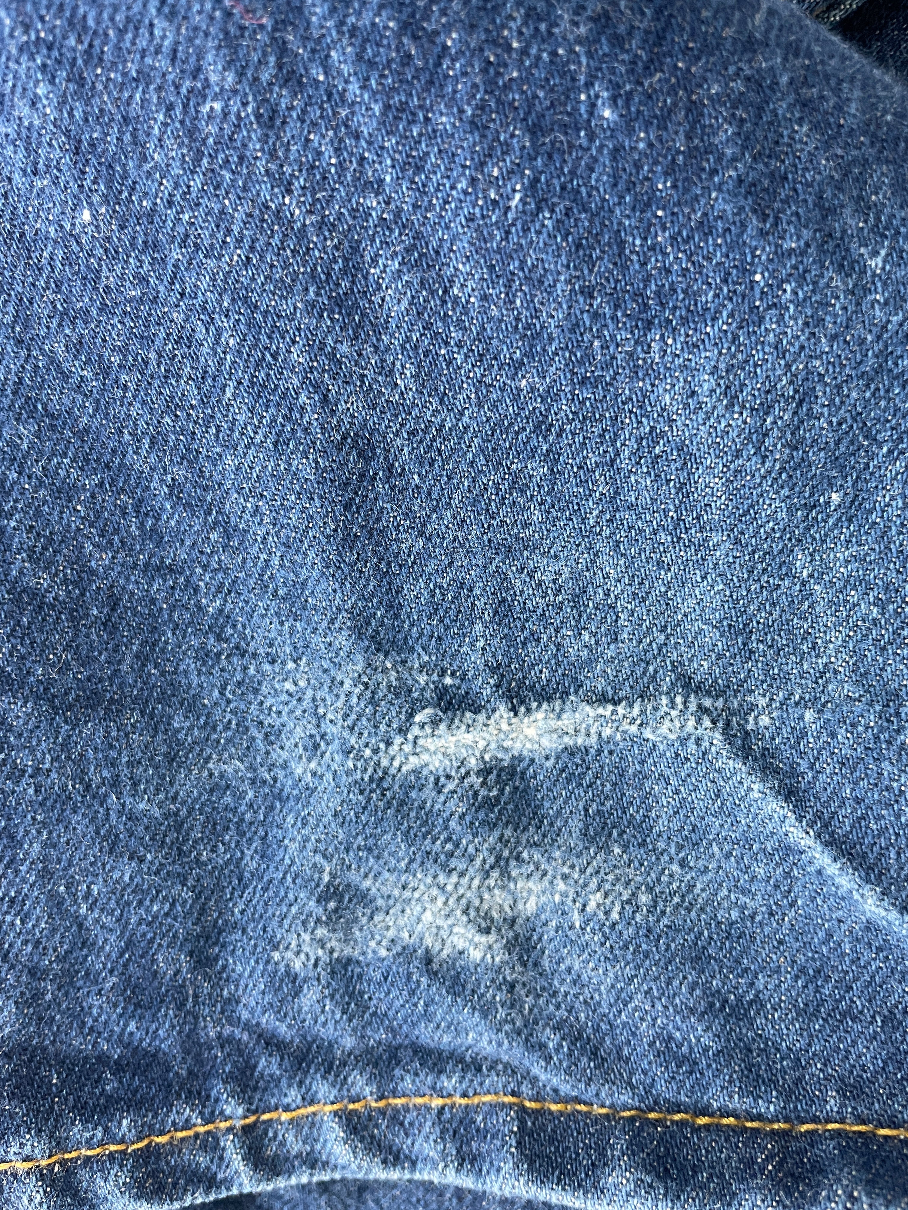 Close-up of blue denim fabric with a visible white stain or scuff and a yellow contrast stitch along the right edge.