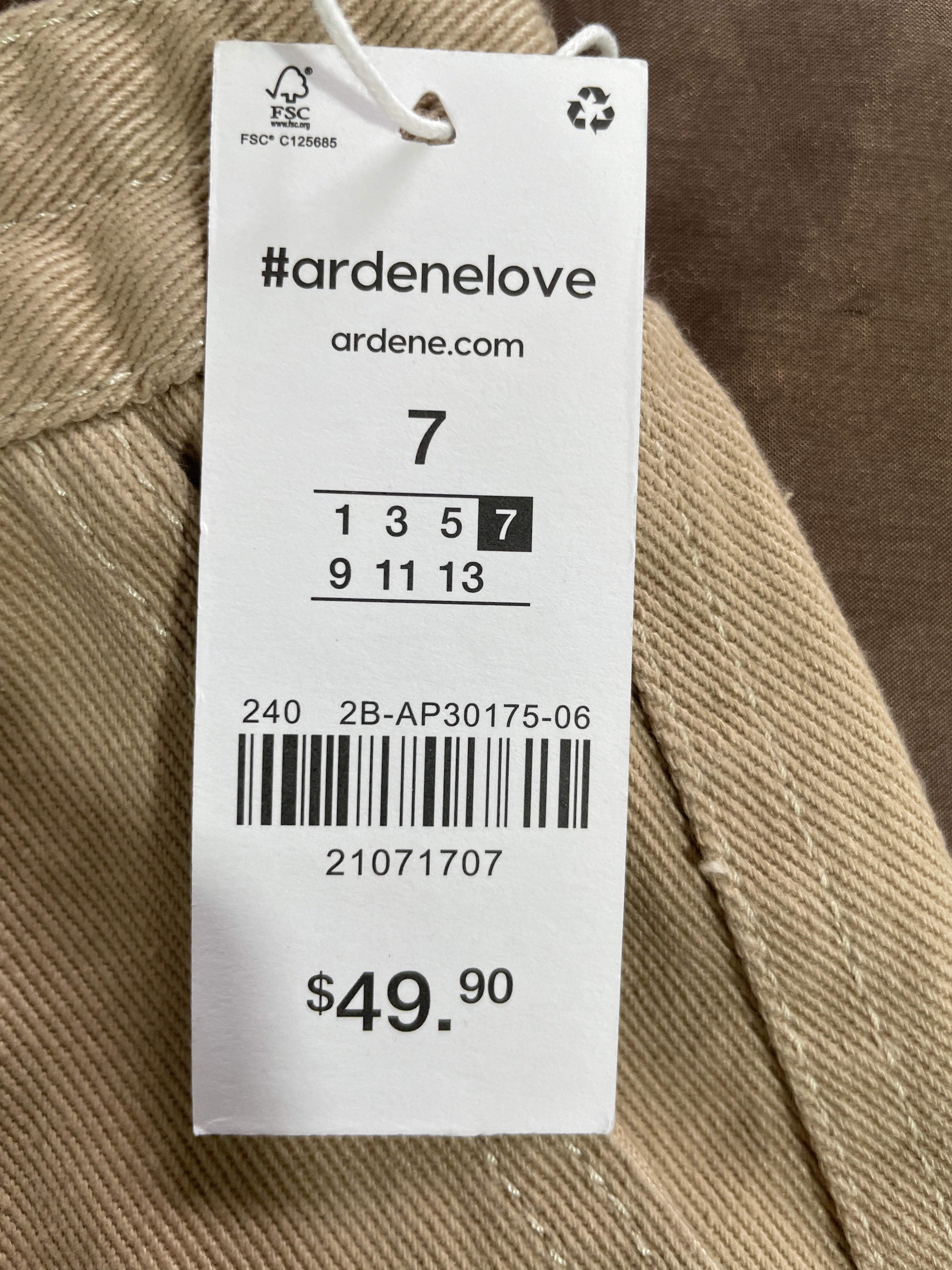 A white Arden Love garment tag attached to a beige woven fabric, showing the brand name, size information, barcode, and price $49.90.