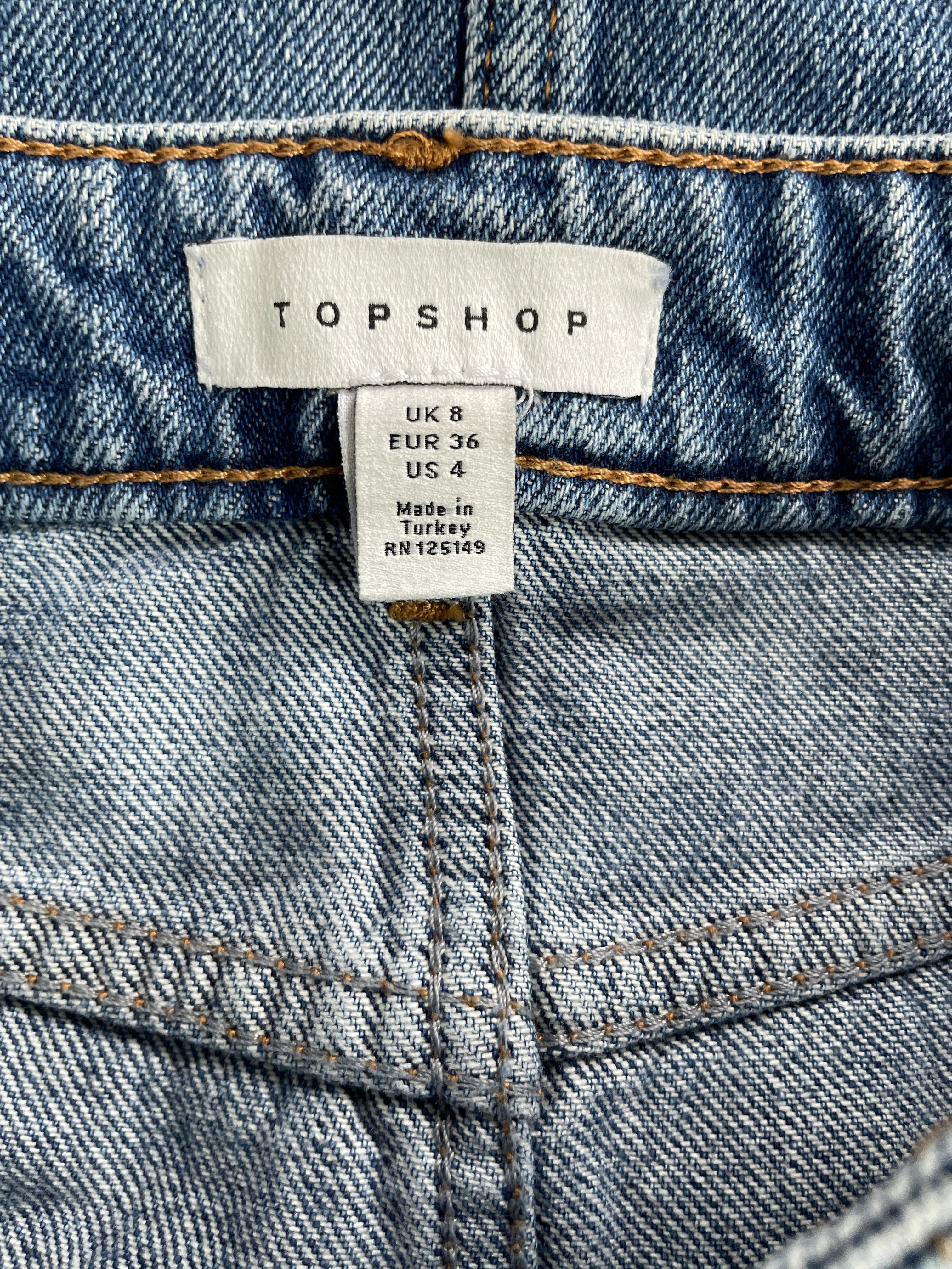 Close-up of a blue denim waistband with a visible TOPSHOP brand label and size text, showing stitched seam details.