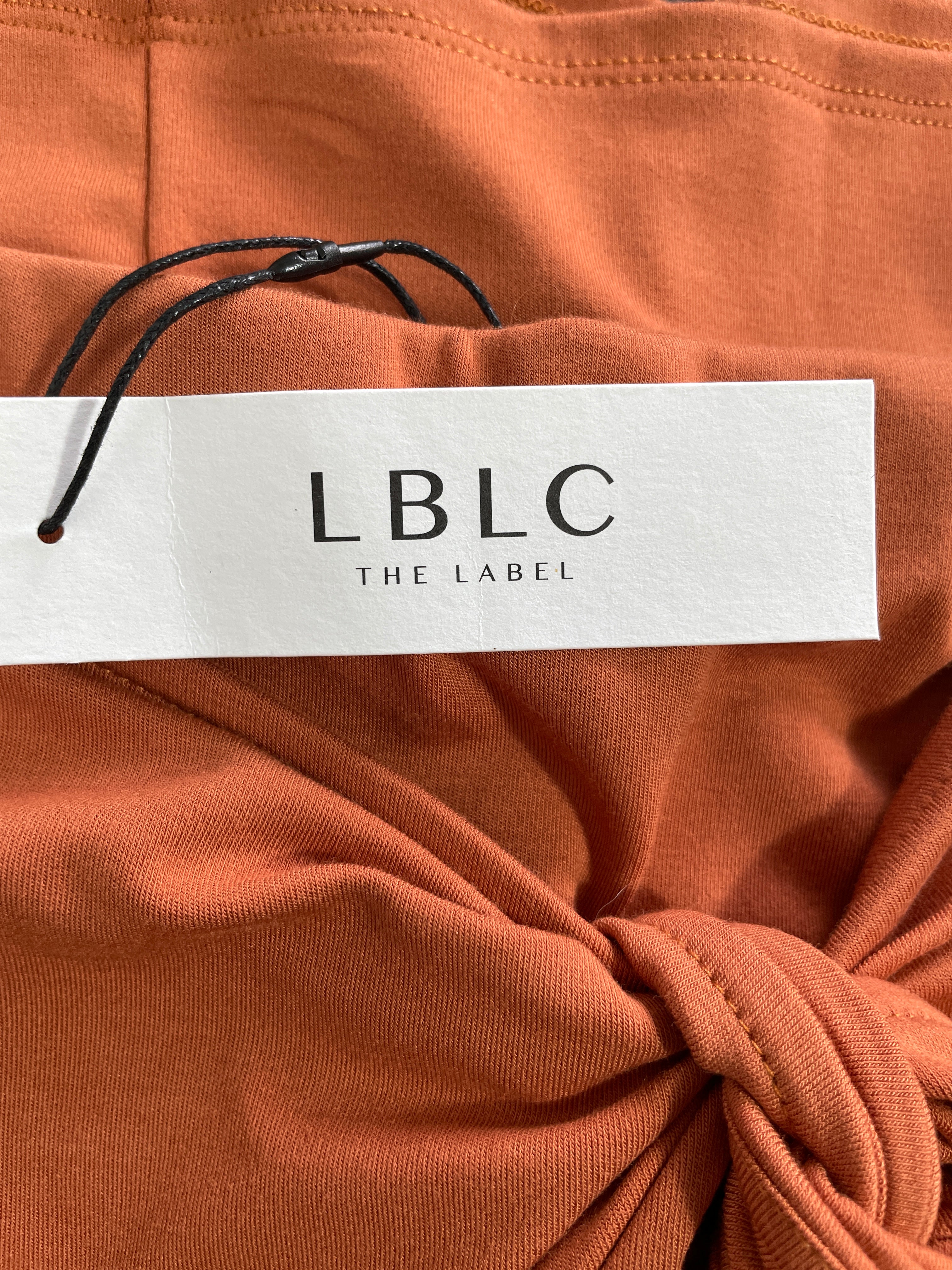 Close-up of rust-orange knit fabric with a knotted tie detail and a white LBLC THE LABEL paper tag attached by a black string.
