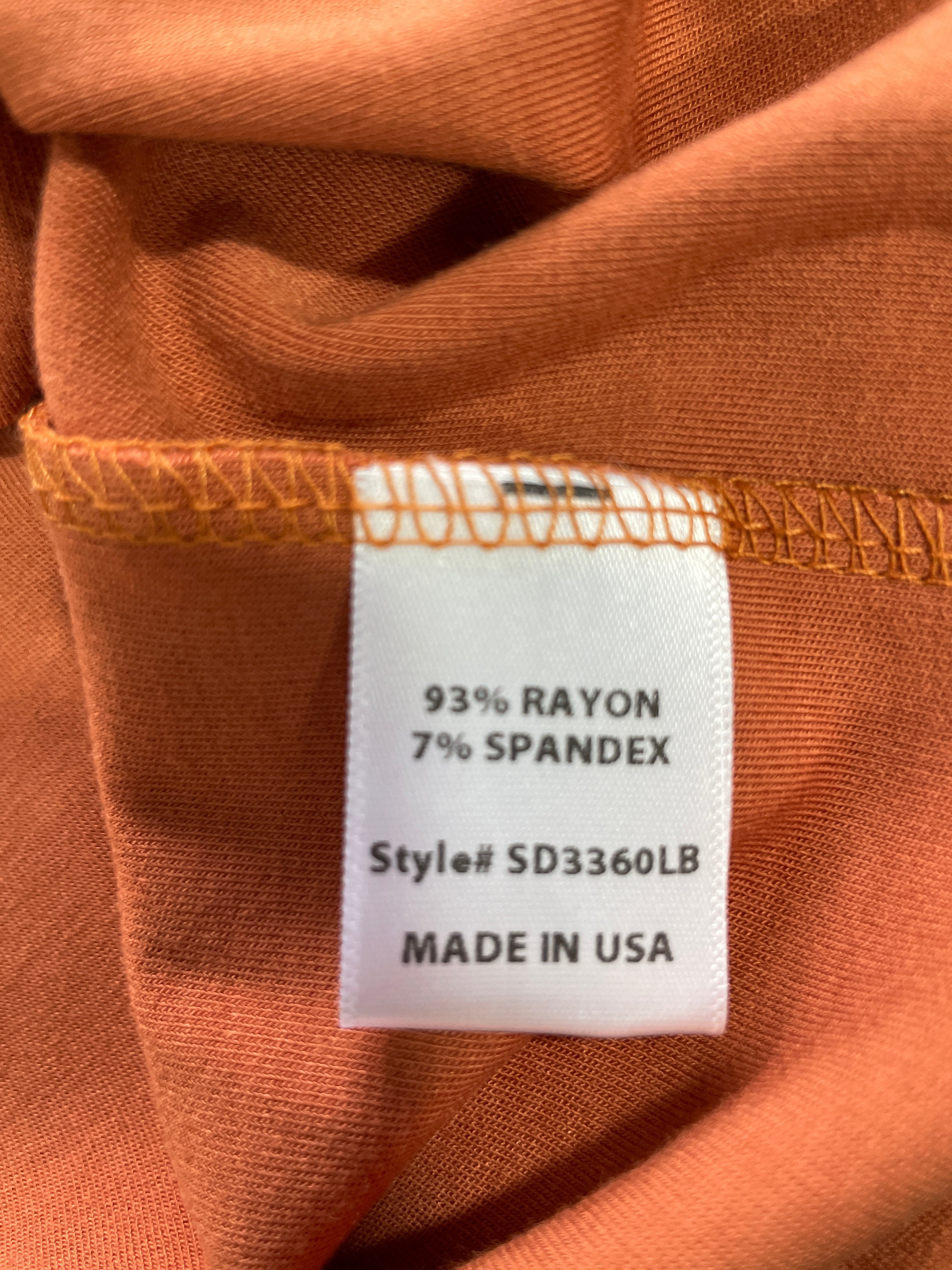 Close-up of a white fabric care label sewn onto an orange-brown garment, showing text that reads '93% RAYON 7% SPANDEX Style# SD3360LB MADE IN USA'.