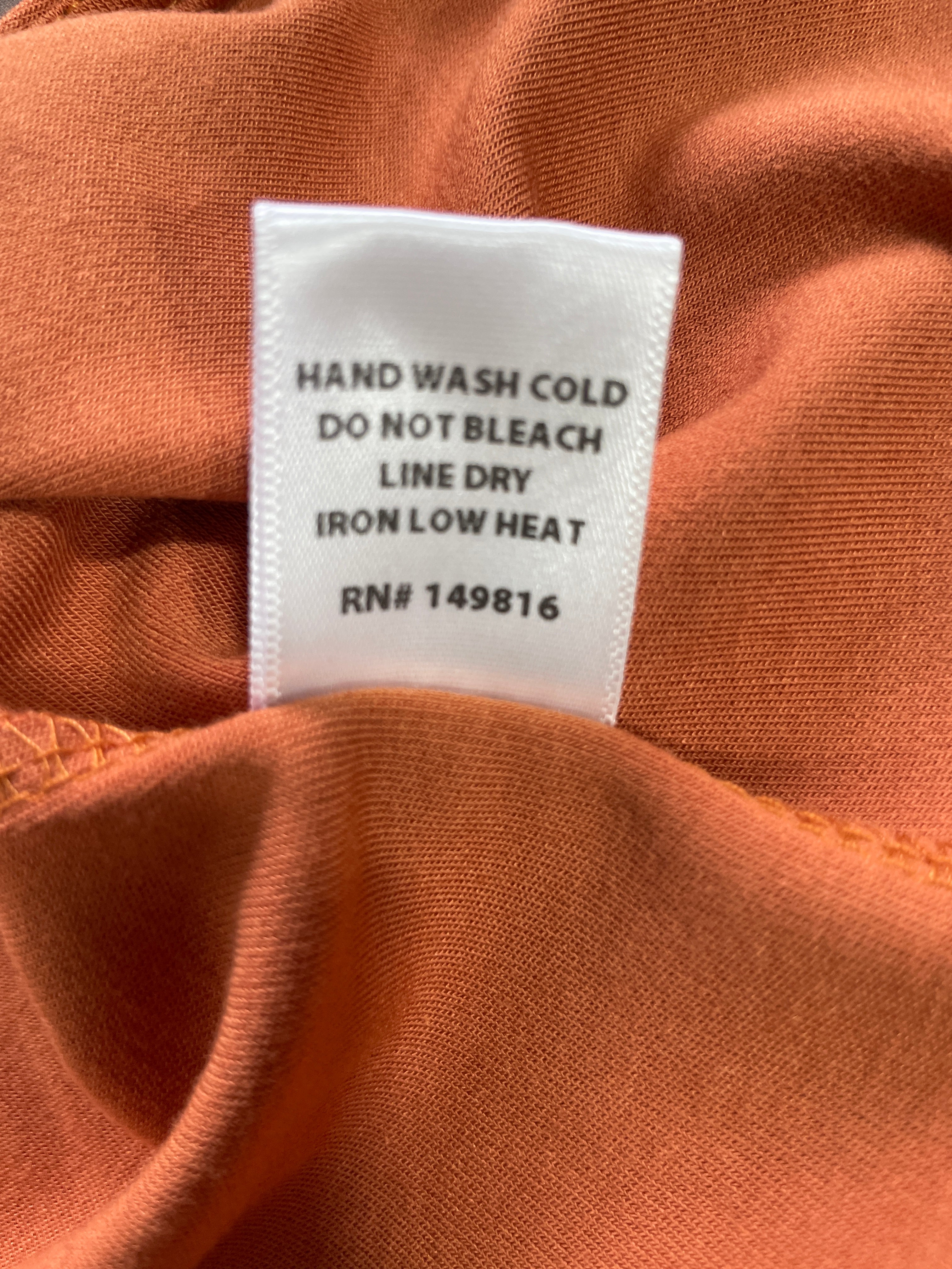 Close-up of a white garment care label sewn onto rust-orange fabric, with printed instructions reading 'HAND WASH COLD', 'DO NOT BLEACH', 'LINE DRY', and 'IRON LOW HEAT'.