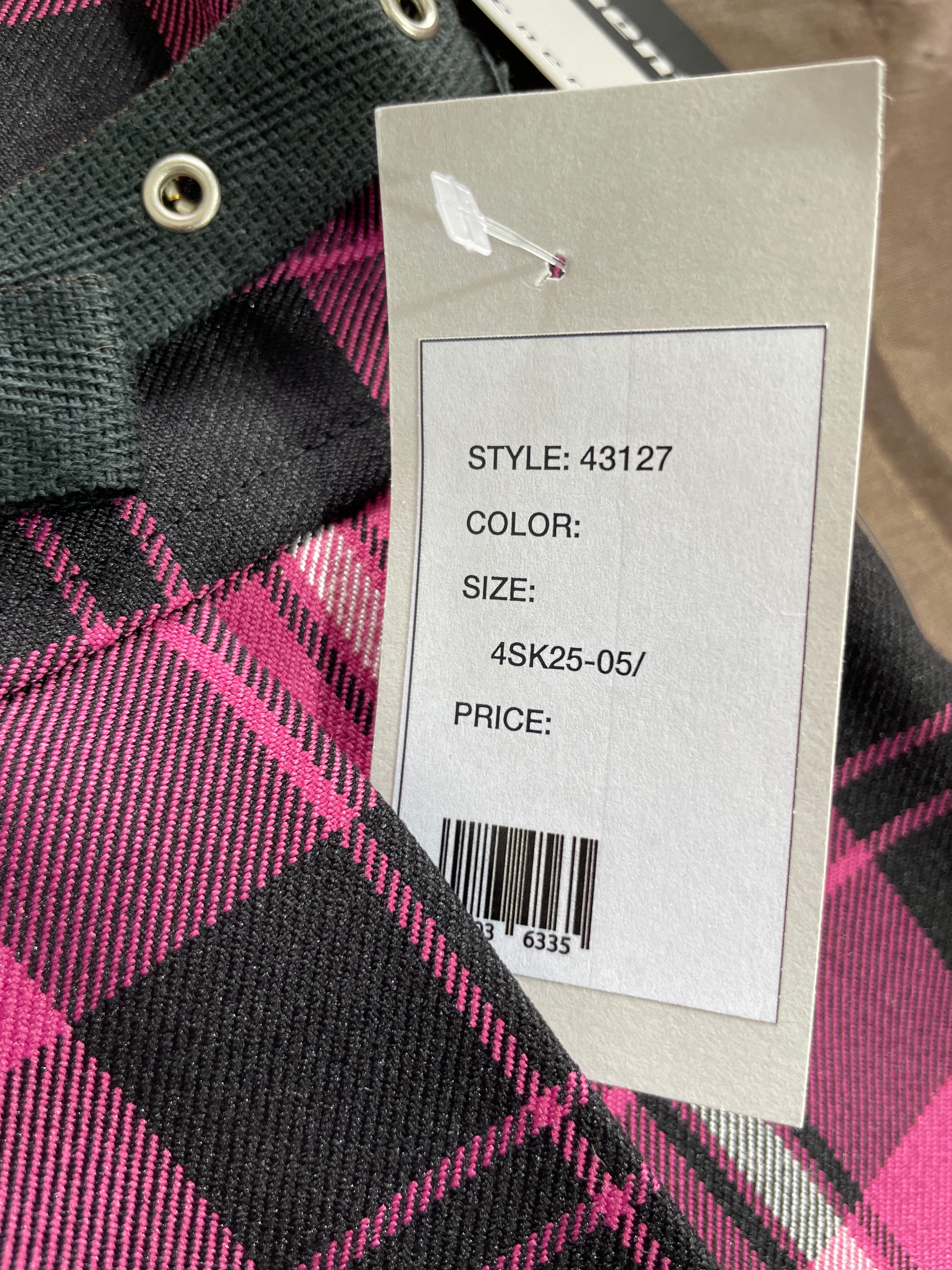 A close-up of a plaid garment in black and bright pink with a white paper hangtag attached, showing printed style and size information.