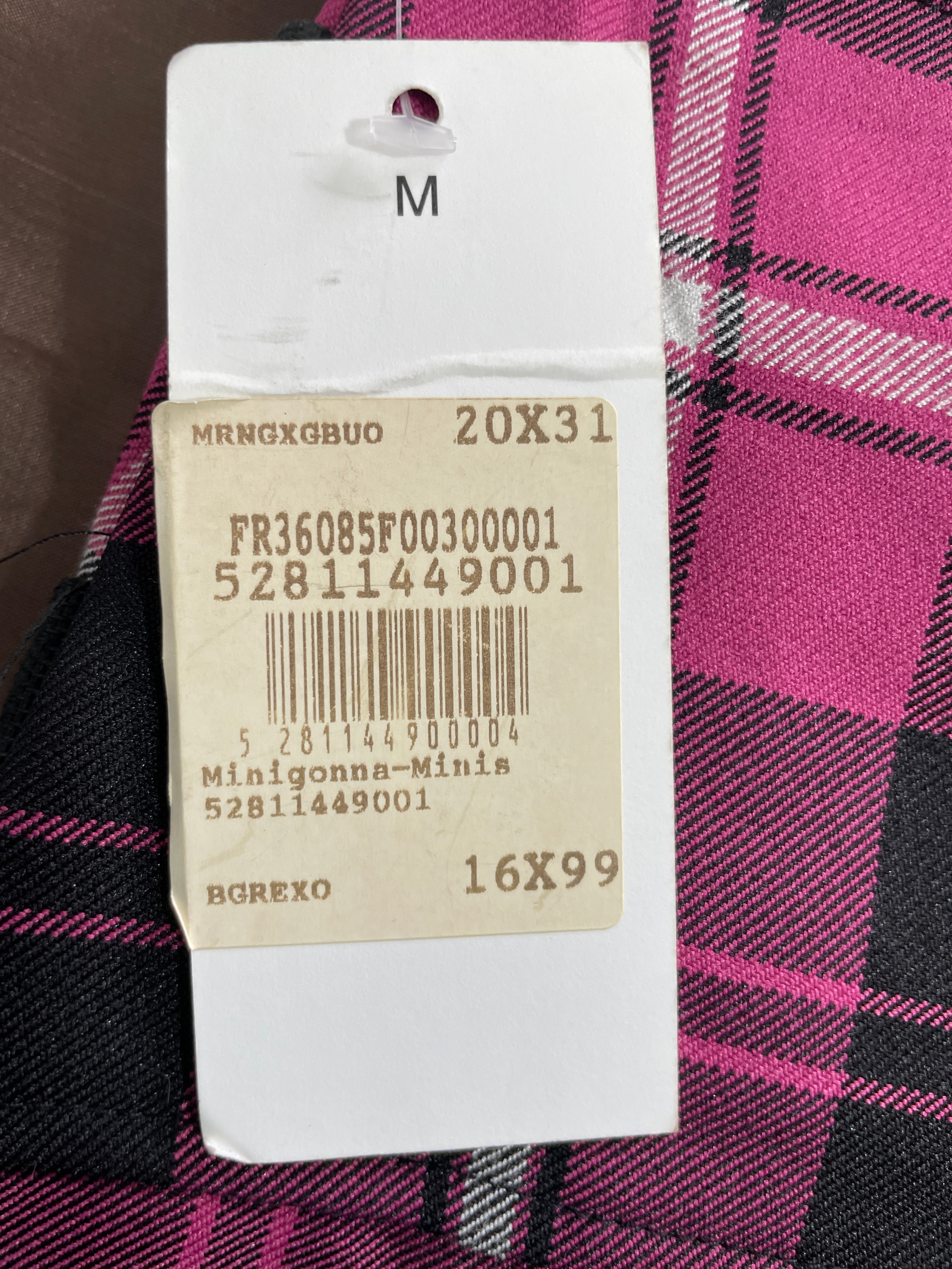 A white clothing size tag with a beige barcode label, placed over a pink, black, and gray plaid fabric. The tag shows text including 'M', '20X31', and 'Minigonna-Minis'.