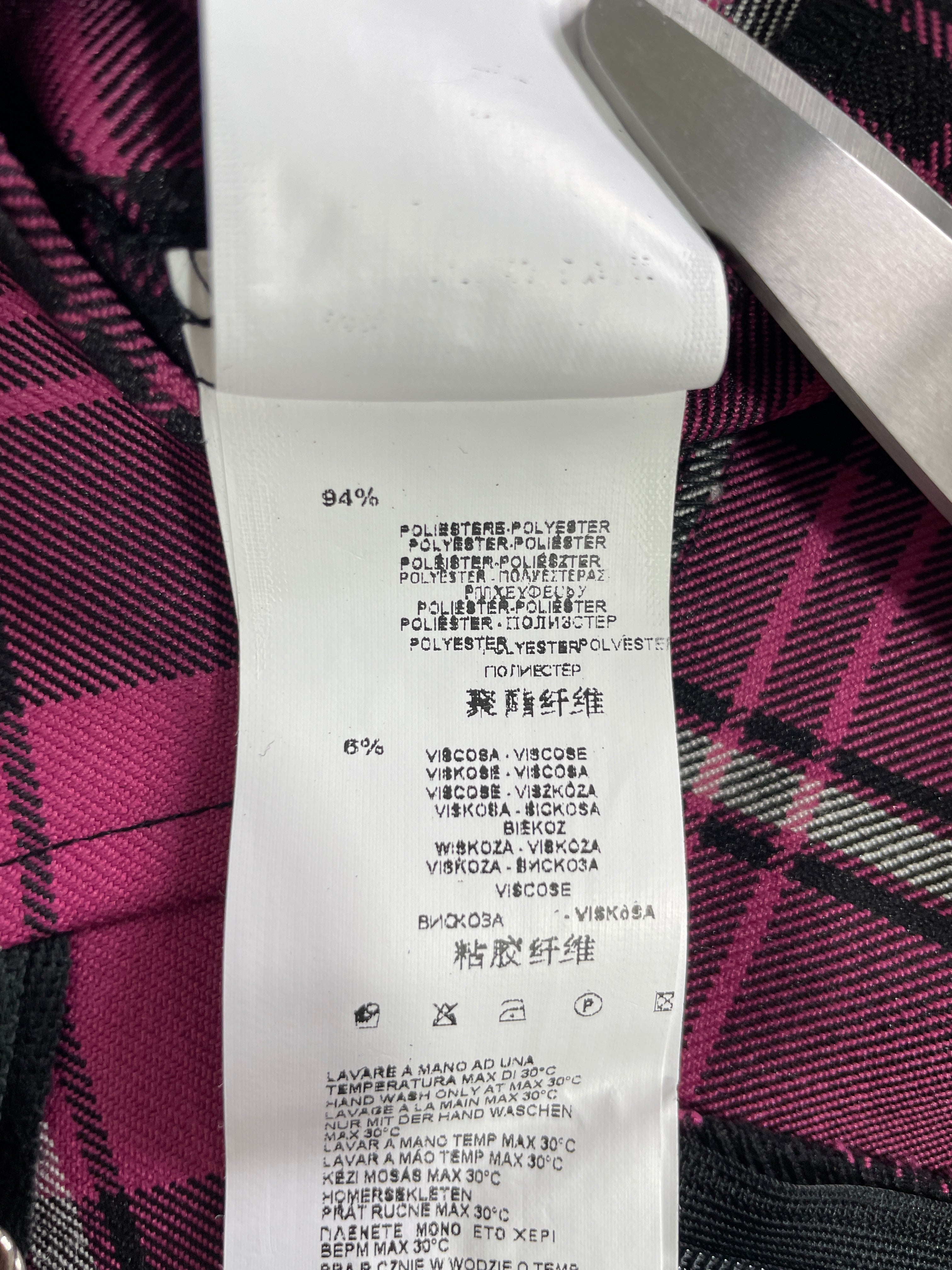 Close-up of a white garment care label sewn onto a pink, black, and gray plaid fabric, with a metal scissors blade visible at the top right.