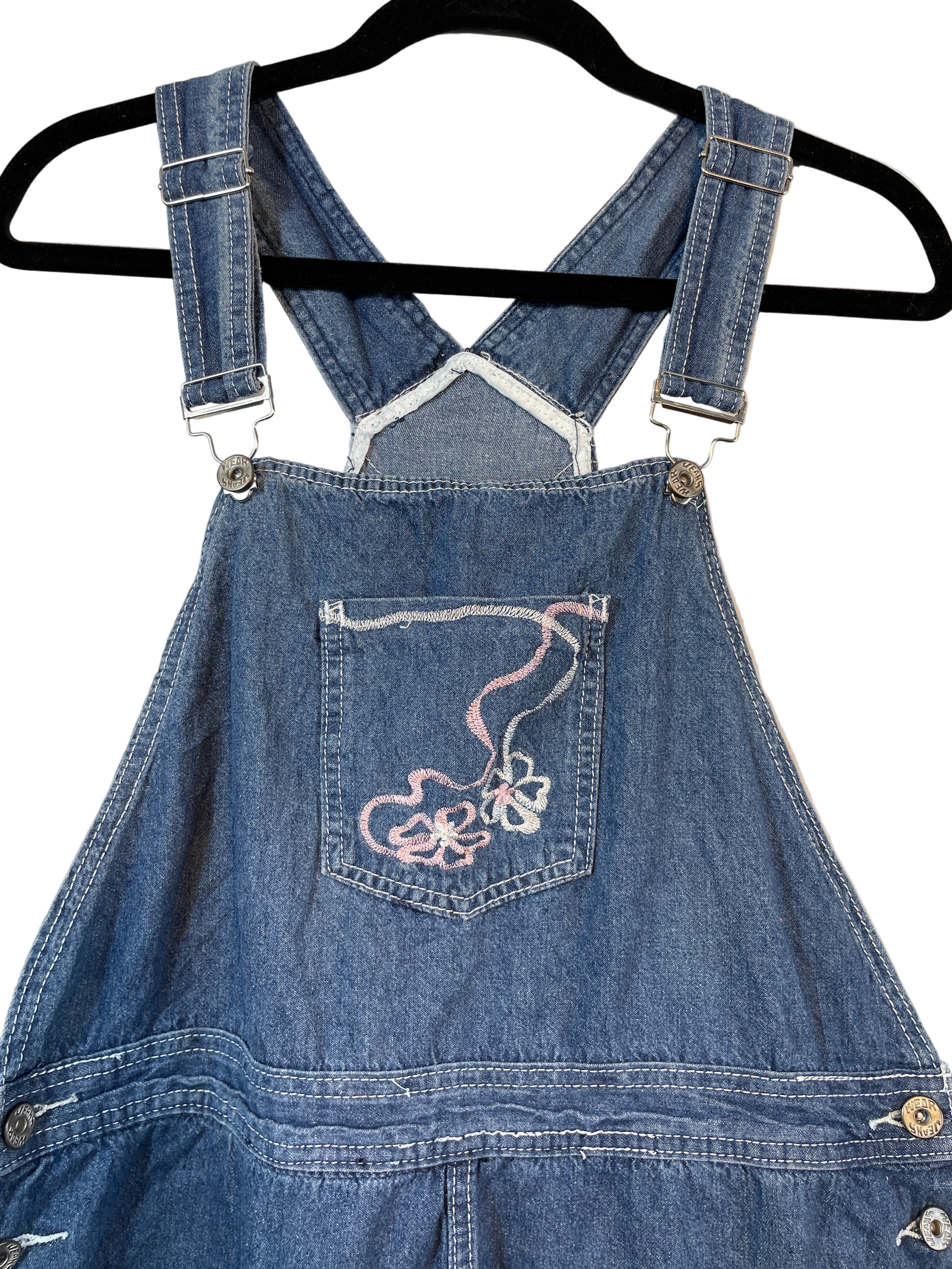 Close-up of PANO denim overalls showing pink and white embroidered flowers on front pocket