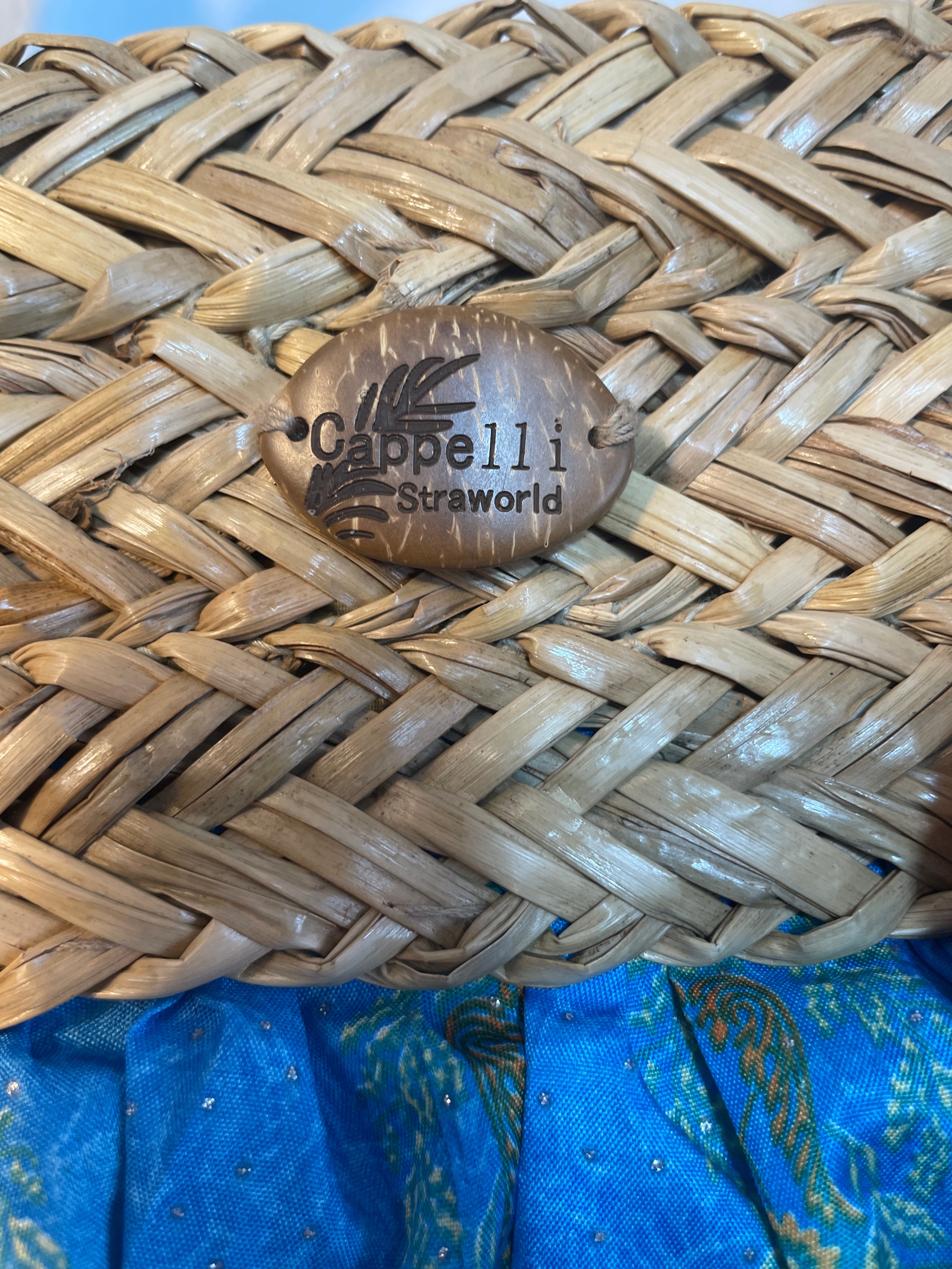 Close-up of a woven straw purse with a round wooden brand plaque reading 'Cappelli Strawworld' in the center, resting on a blue patterned fabric surface.
