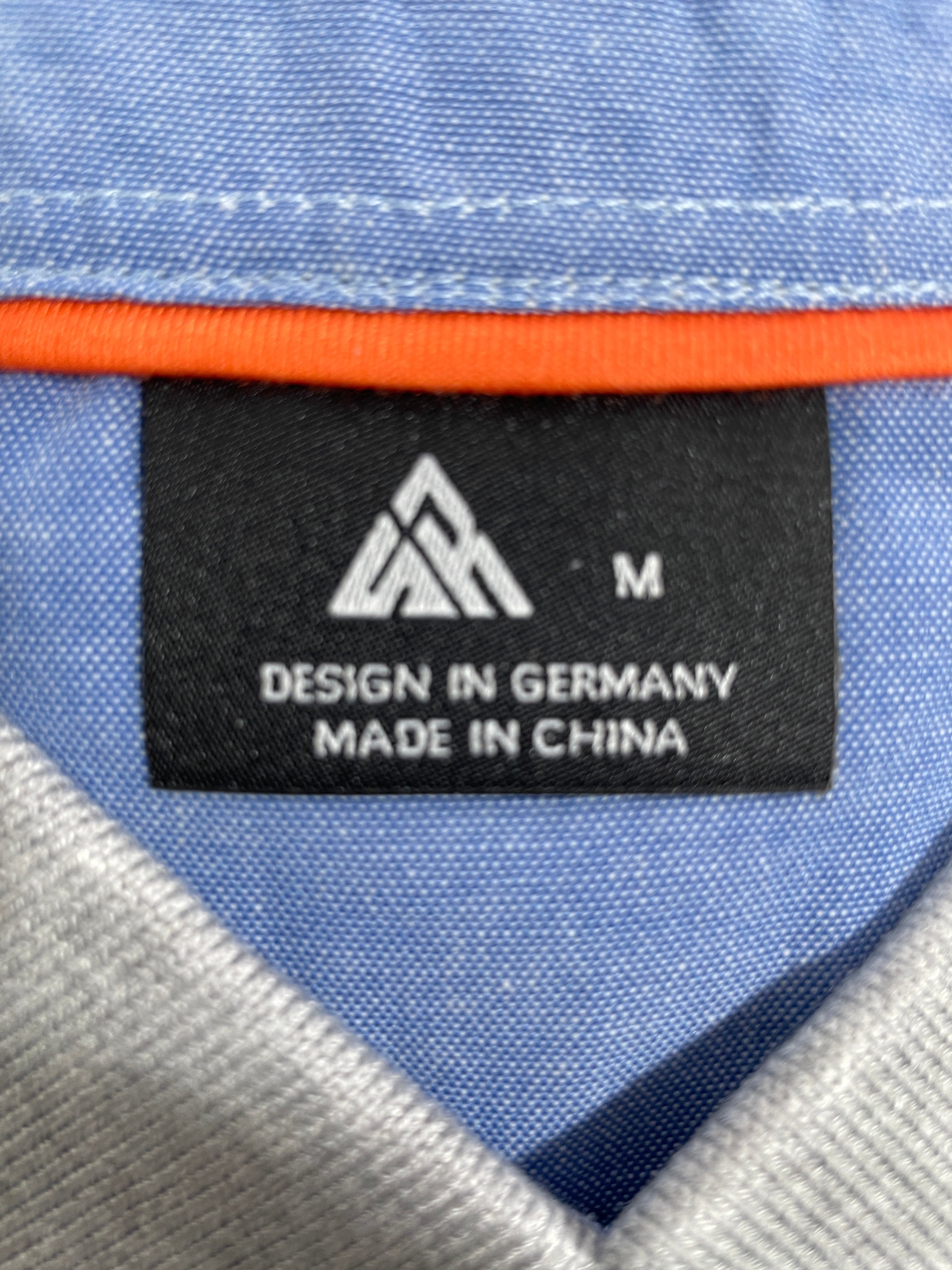 Close-up of a garment interior label reading 'DESIGN IN GERMANY MADE IN CHINA' with a white geometric logo on a black patch, surrounded by light blue fabric and an orange trim.