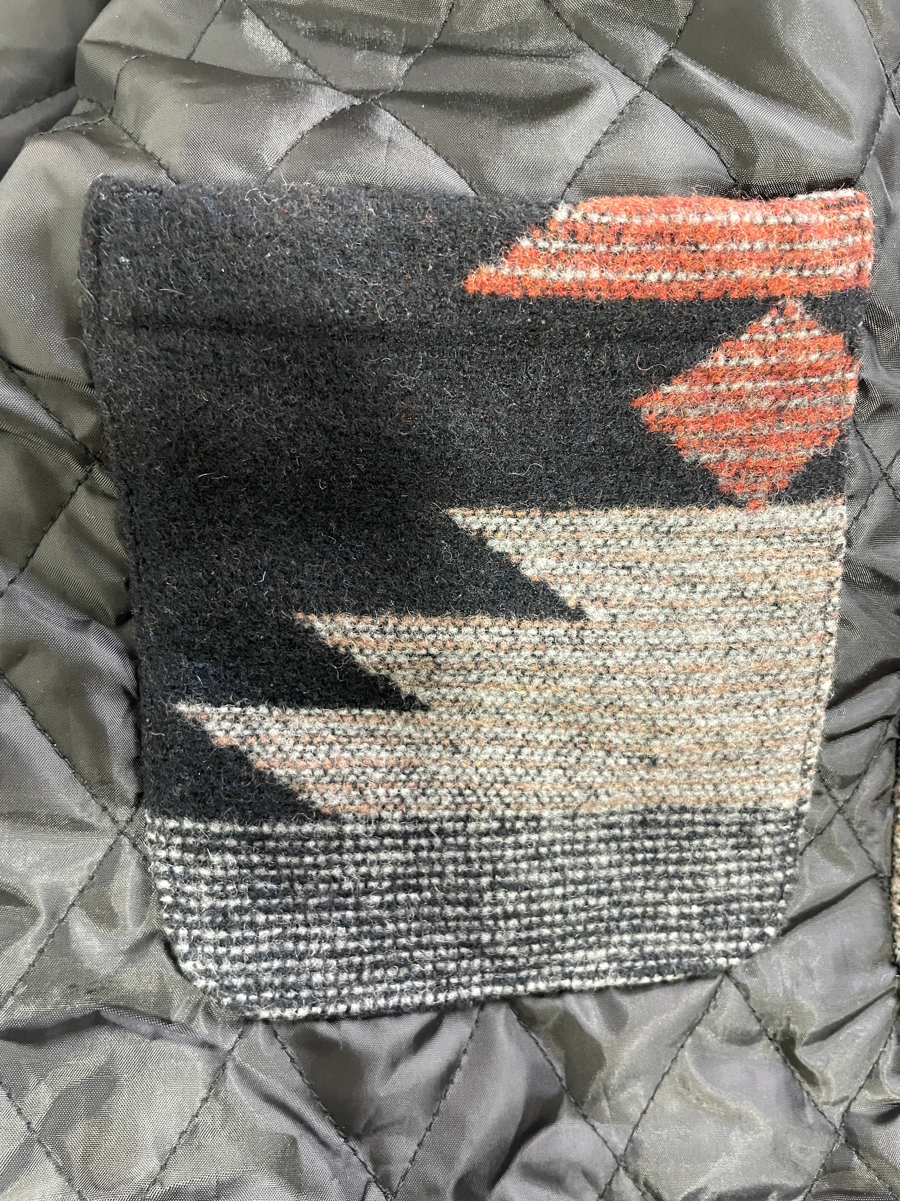 A close-up of a woven textile patch with a Southwestern geometric pattern in black, beige, gray, and terracotta orange, laid flat on a gray quilted background.
