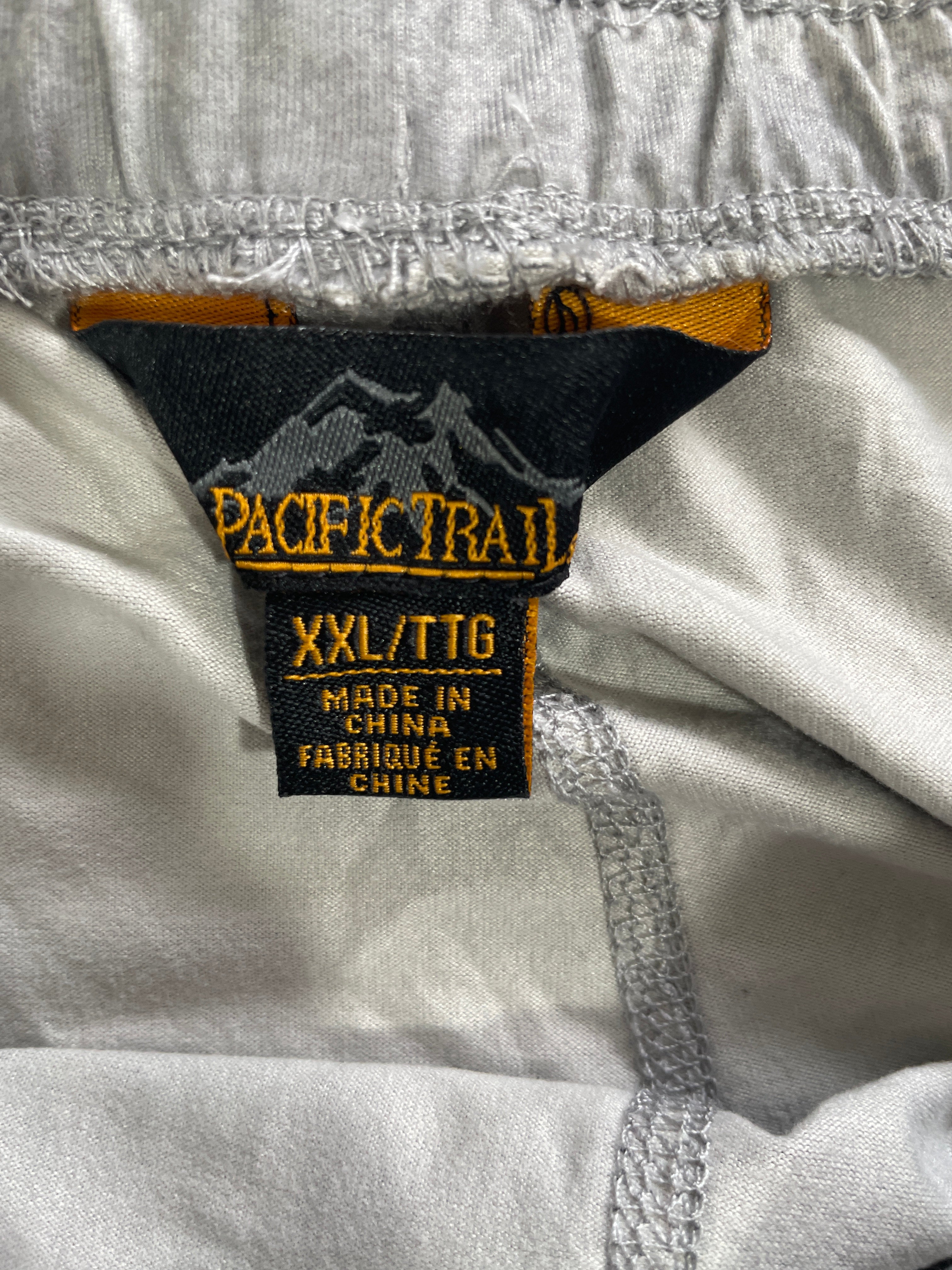 Close-up of a garment's interior neck label reading PACIFIC TRAIL, size XXL/TTG, with a black and orange brand tag stitched onto light gray fabric.