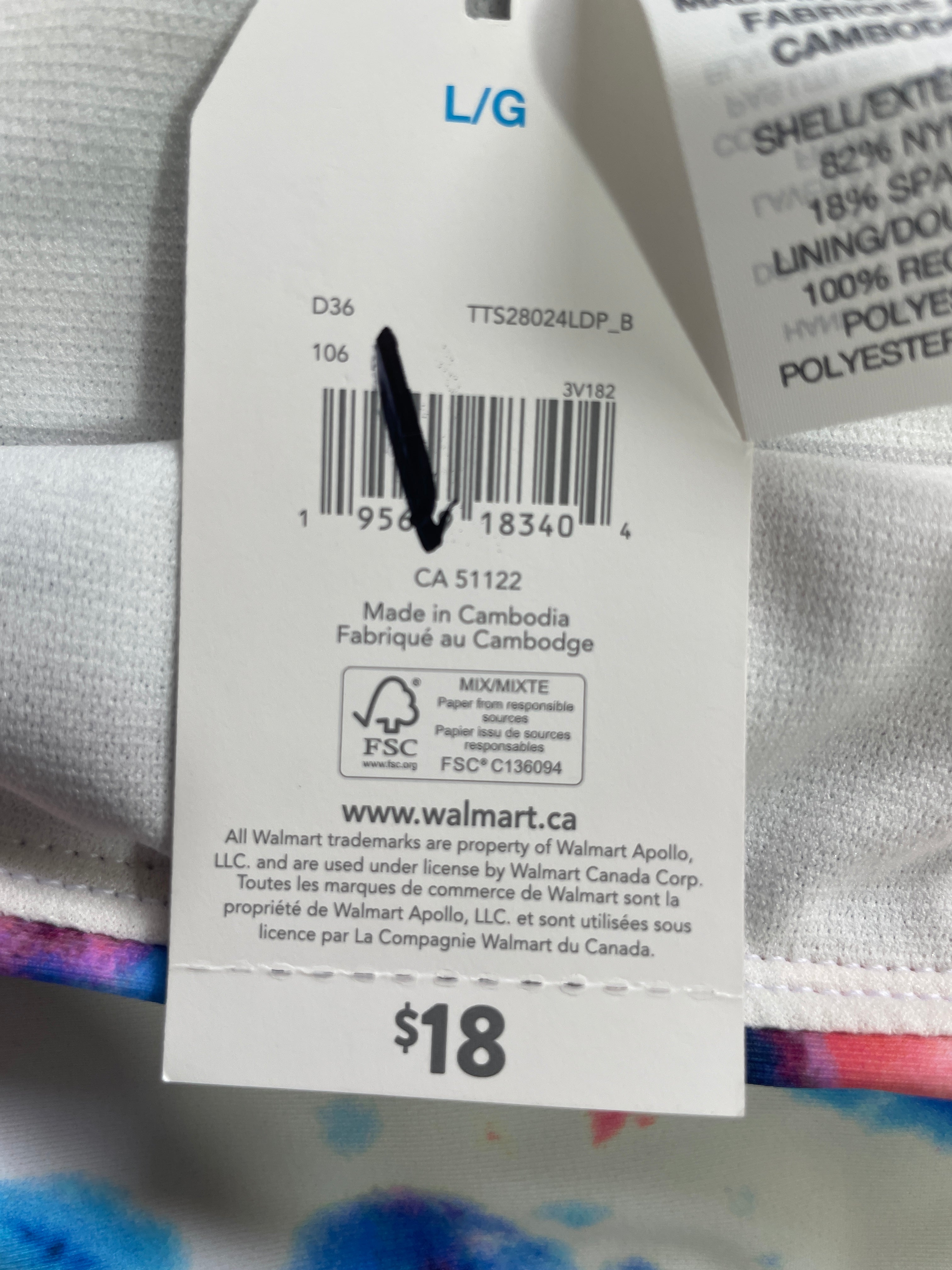 A close-up of a white garment tag attached to a folded piece of fabric, showing printed details including a barcode, size code D36, Walmart website text, and a price of $18. A black pen points at the barcode.