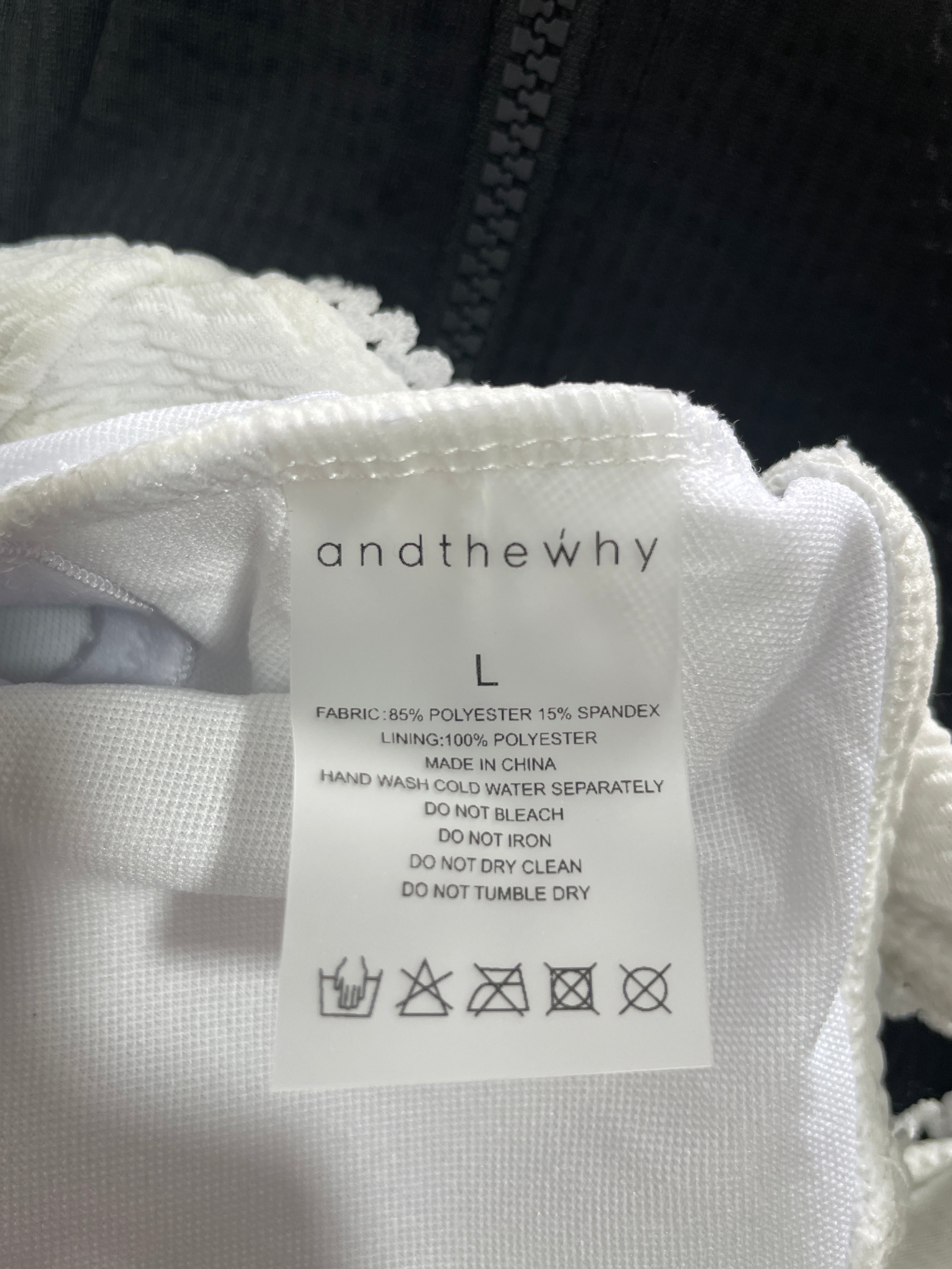 Close-up of a white fabric garment tag reading 'and the why' and size 'L', attached to a white fringed textile, with a dark zipper or fabric visible in the background.