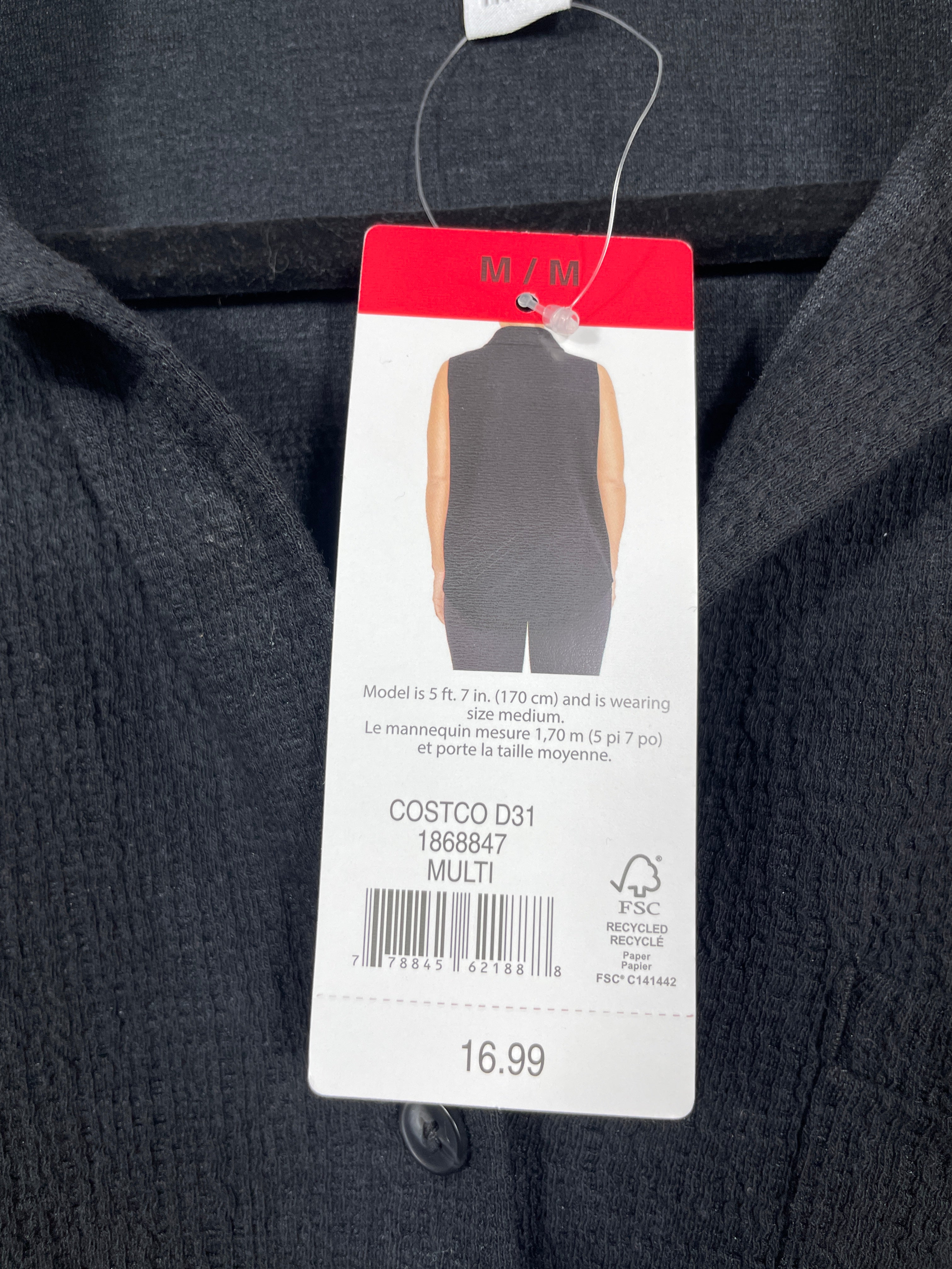A close-up of a dark charcoal gray knit garment with a retail hangtag attached. The tag shows a model wearing a sleeveless vest and includes text reading 'COSTCO D31', '1268947', 'MULTI', and the price '16.