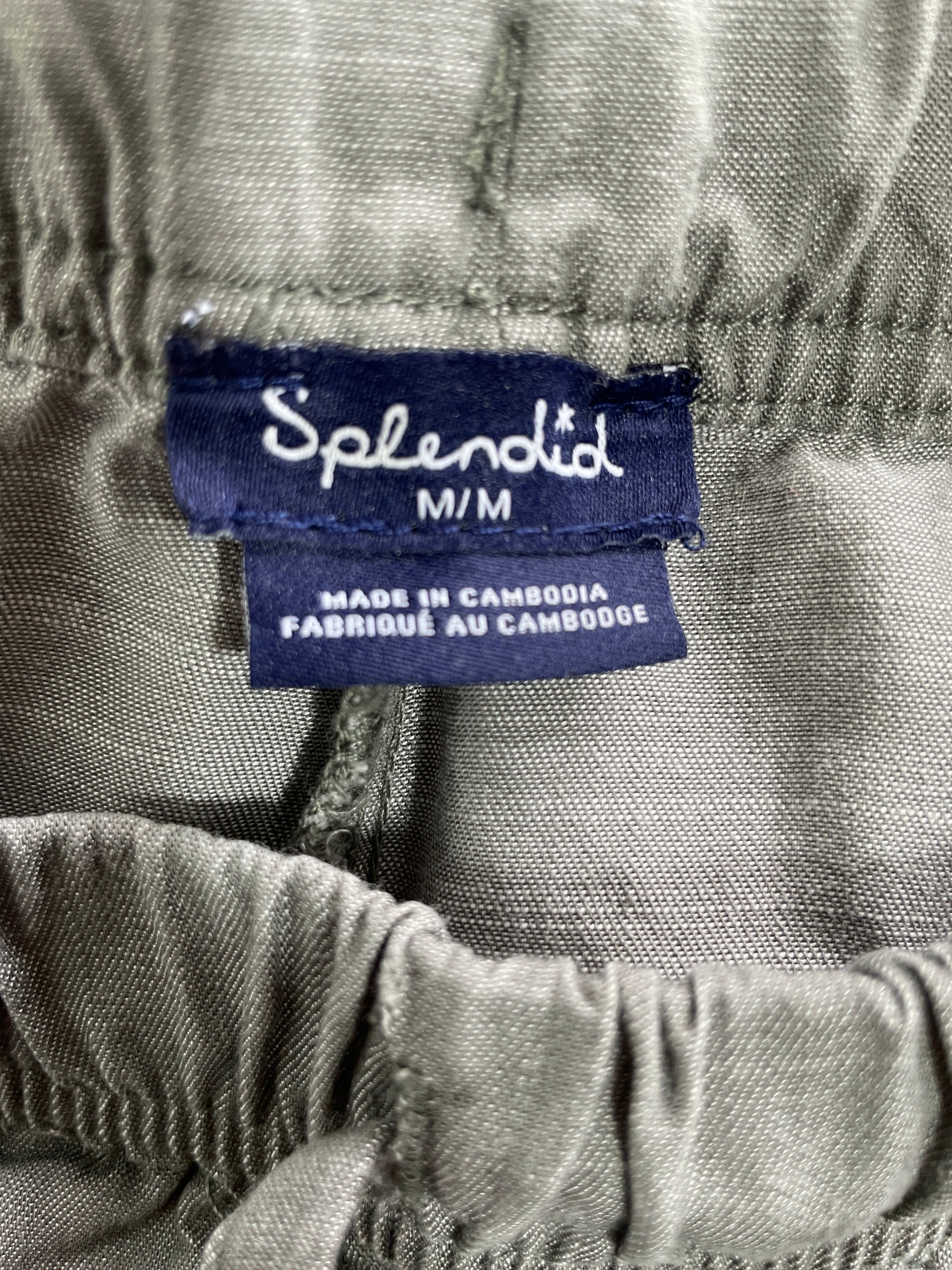Close-up of a garment label reading 'Splendid M/M' and 'MADE IN CAMBODIA' on a gray fabric, with gathered elastic trim visible around the waistband.