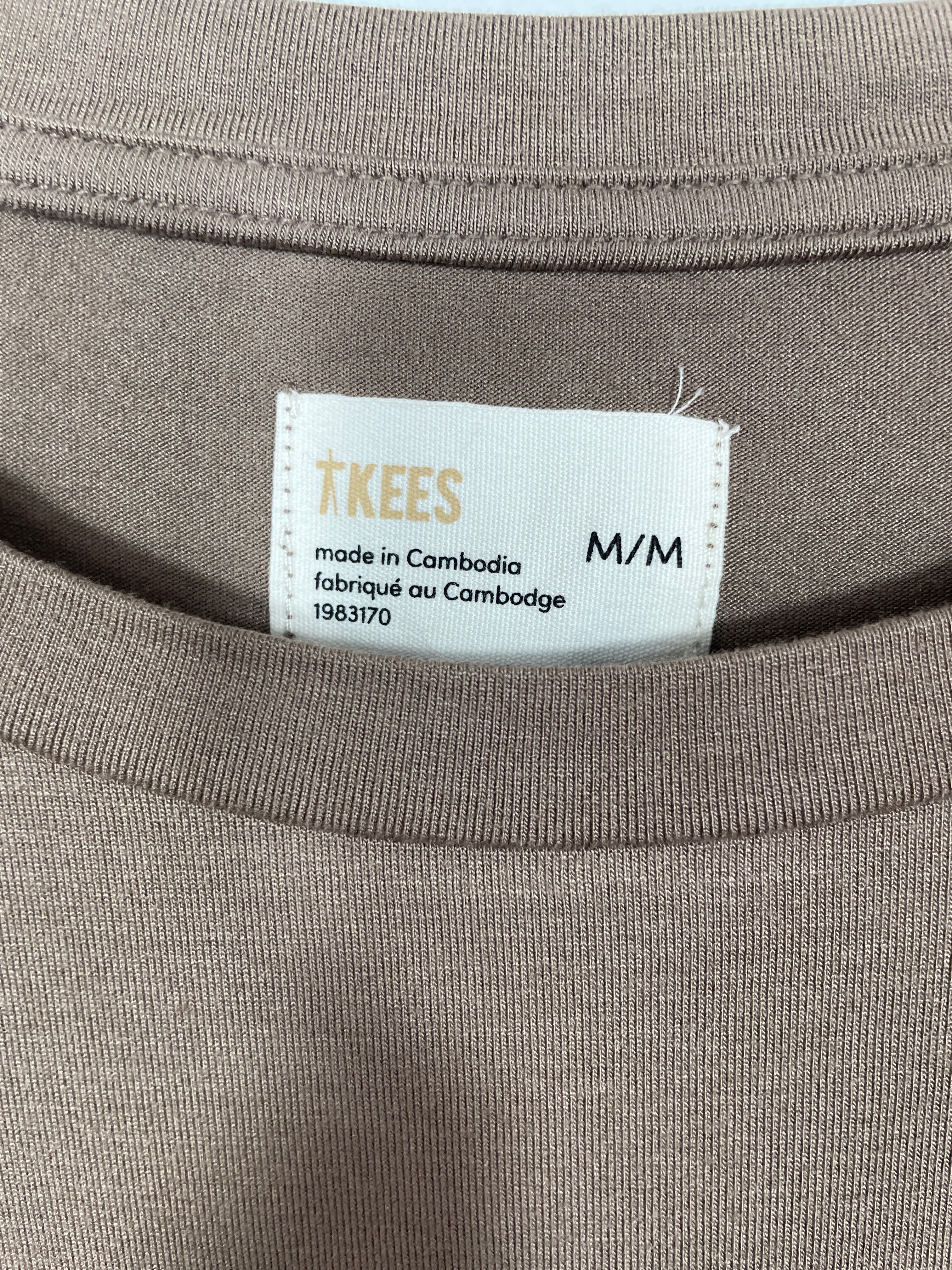 Close-up of a taupe-brown t-shirt neck label reading 'TKEES' and 'M/M', with ribbed collar and fabric texture visible.