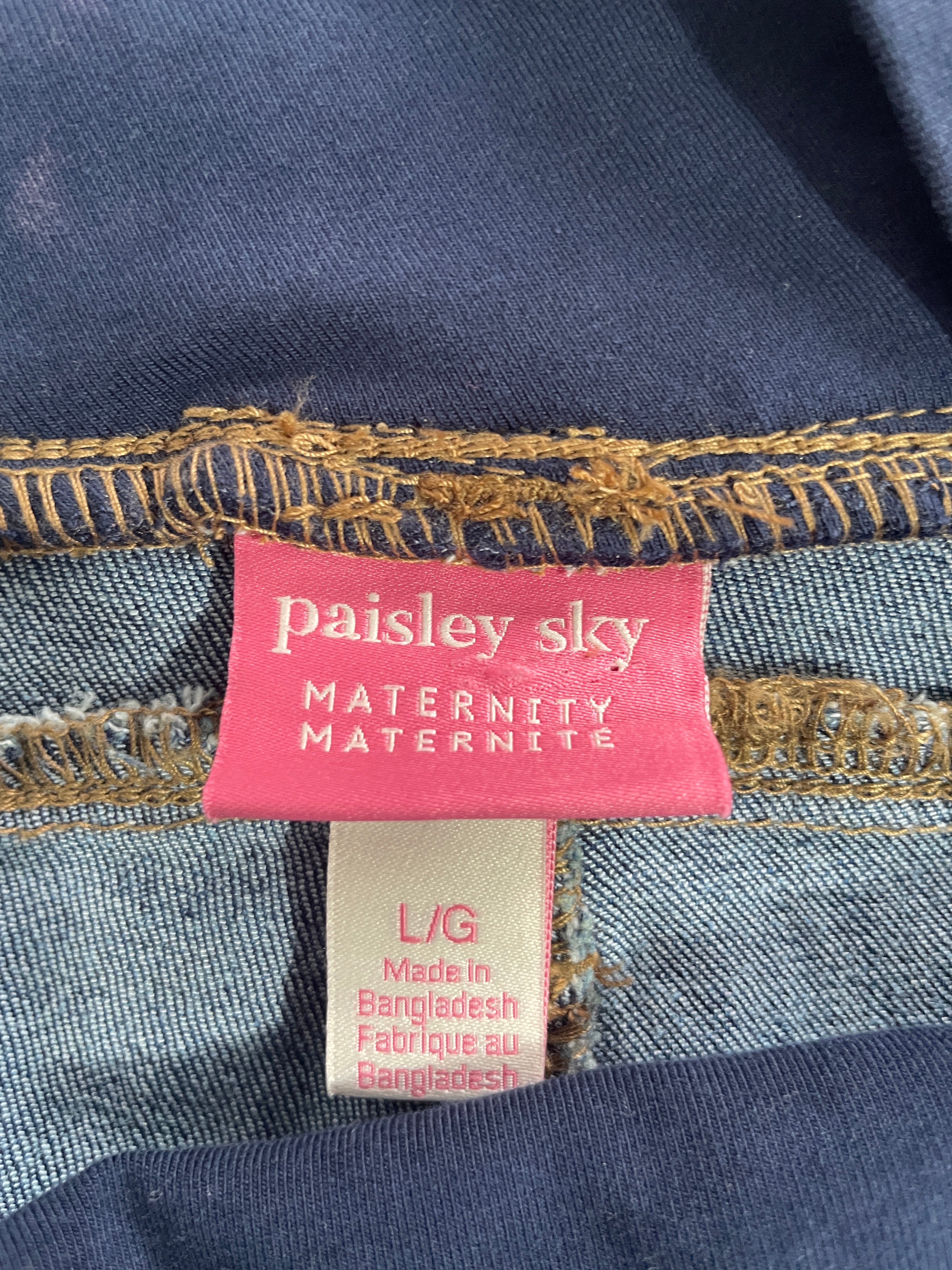 Close-up of a denim maternity garment showing a pink brand label that reads 'paisley sky MATERNITY MATERNITE' and a beige size tag reading 'L/G Made in Bangladesh'.