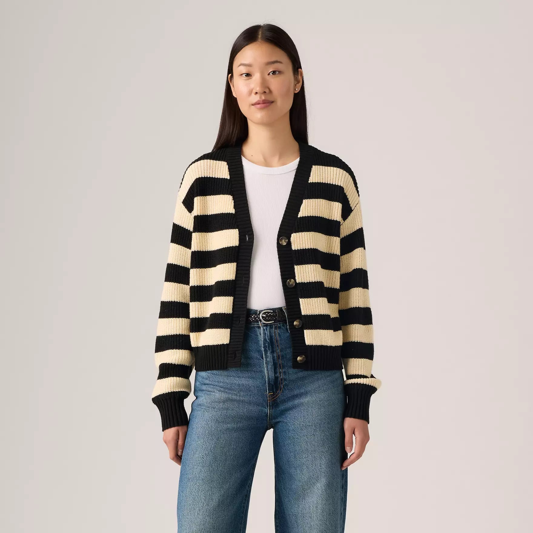 Woman wearing a black and beige striped cardigan with blue jeans on a plain background