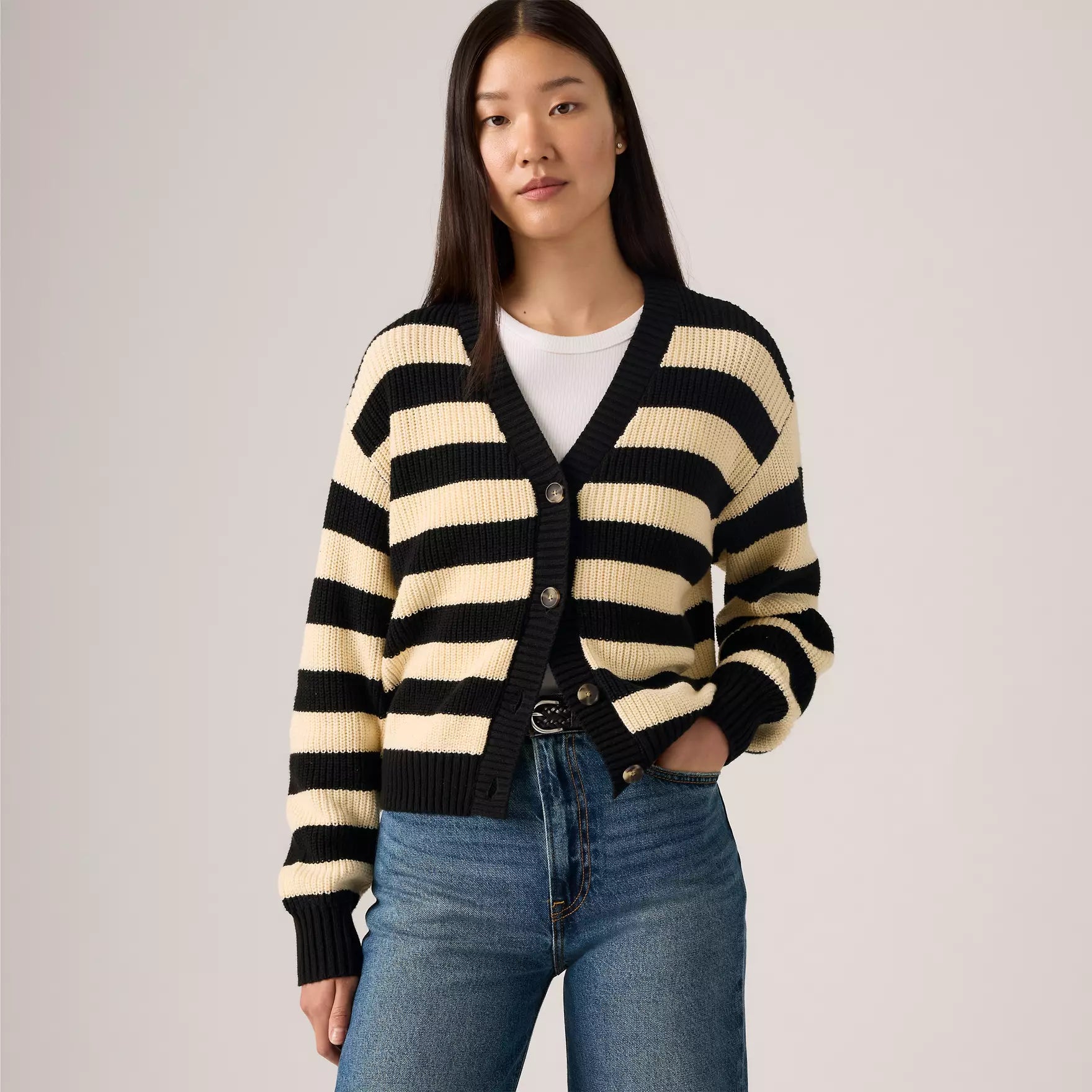 Woman wearing a black and beige striped cardigan with blue jeans on a plain background